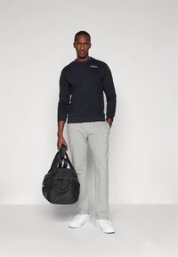 Conjunto deportivo que incluye una sudadera azul marino, pantalones de chándal grises y zapatillas blancas. Sosteniendo una bolsa de viaje negra con superficie texturizada.