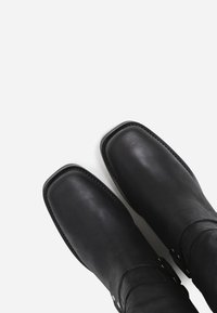 Botas de tobillo de cuero negro con punta cuadrada, textura suave y herrajes decorativos en el lateral. Detalles de costura a lo largo de los bordes.