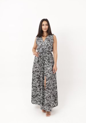 Femme portant une robe maxi noire et blanche à motifs floraux sans manches avec une fente sur le devant, debout devant un fond blanc uni.