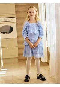 Abito azzurro chiaro con maniche a sbuffo e volant, abbinato a collant bianchi e scarpe nere. Tessuto liscio con una leggera consistenza.