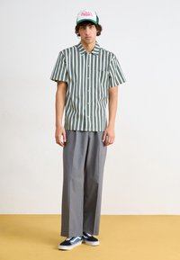 Chemise à manches courtes avec col, rayures verticales vertes et blanches, boutonnage devant, portée avec un pantalon gris ample et des baskets bleues.