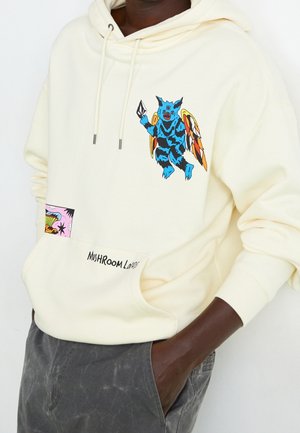 Sweat à capuche crème en tissu doux, ornée d'un graphique coloré d'une créature bleue avec des ailes, et d'une poche avec un design rose et noir, étiquetée "MUSHROOM."