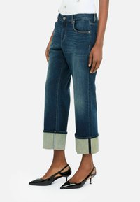 Jeans de mezclilla azul oscuro con dobladillos remangados; presentan costuras en contraste y un diseño clásico de cinco bolsillos. Llevados con tacones negros de punta.