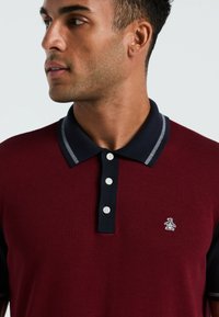 Bordeaux polo shirt met marineblauwe kraag en afwerking, voorzien van drie witte knopen en een geborduurd pinguïnlogo op de linkerborst.