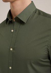 Zbliżenie na osobę w oliwkowej koszuli zapinanej na guziki z kołnierzykiem zakończonym w szpic, na neutralnym tle.