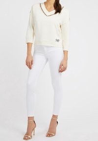 Pullover bianco lavorato a maglia con scollatura a V e maniche corte. Presenta dettagli a catena sul collo e un logo nella parte inferiore. Abbinato a jeans skinny bianchi.