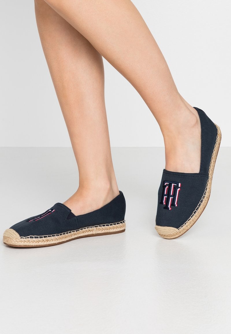 Jambes portant des espadrilles plates noires avec des semelles beiges tressées et un logo brodé rouge-blanc-bleu sur le dessus, debout sur un sol blanc.