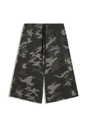 Ung kvinde med krøllet hår iført hvid og rød cropped t-shirt, grå camouflage vidde shorts og sorte flip-flops står ved et spejl udendørs.