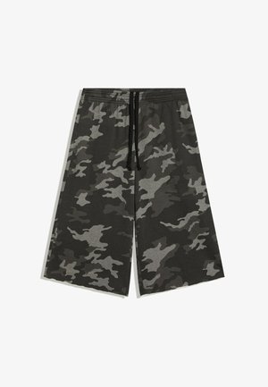 Junge Frau mit lockigem Haar trägt ein weißes und rotes kurzes T-Shirt, graue Camouflage-Weitbein-Shorts und schwarze Flip-Flops und steht draußen an einem Spiegel.