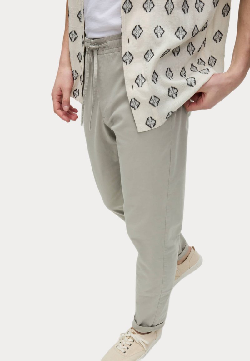 Pantalones grises claros con cintura elástica y cordón, combinados con una camisa crema de estampado. El calzado son unas zapatillas ligeras de color beige.