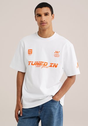 Mladý muž s krátkymi tmavými vlasmi, oblečený v bielom tričku s oranžovým textom "TUNED IN club spirit" a modrých džínsoch, stojaci s jednou rukou v vrecku.