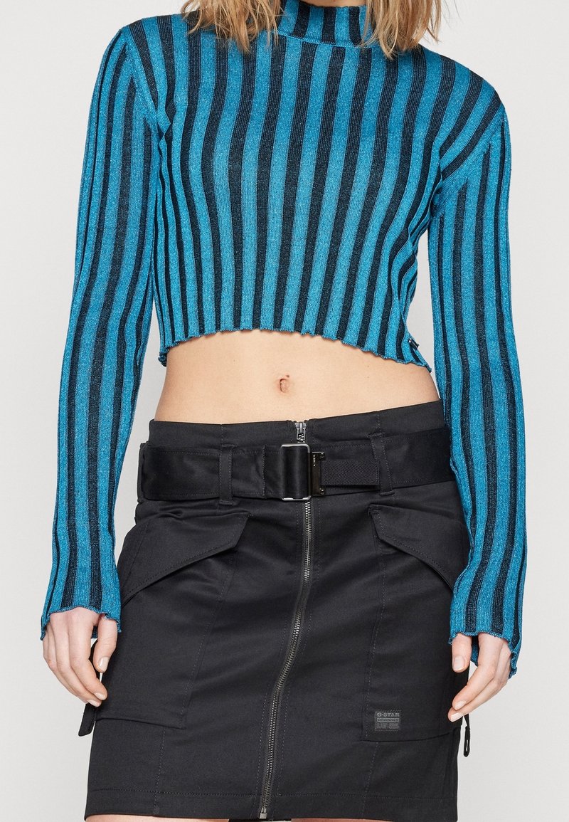 Modrý a černý pruhovaný zkrácený svetr s dlouhými rukávy, který je kombinován s černou sukní s vysokým pasem, obsahující přední zip a páskové akcenty.