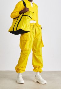 Geel cargo broeken met zijzakken, gecombineerd met een bijpassende lange mouwen top. Een grote gele schoudertas met zwarte accenten maakt de outfit compleet.