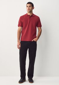 Polo rouge à manches courtes avec col, associé à un pantalon noir droit et des chaussures noires, posé sur un fond uni.