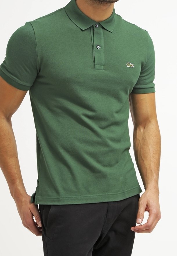 Homme portant un polo vert ajusté à manches courtes avec deux boutons et un petit logo crocodile sur la poitrine, associé à un pantalon noir.