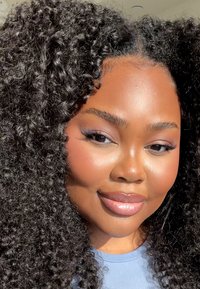 Porträtt av en person med volymöst, lockigt svart hår, jämn hy och en ljuslila skjorta, med mjukt ljus och naturlig makeup.