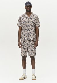 Beige Kurzarm-Set mit blauem Blumenmuster; besteht aus einem Hemd mit Kragen und Shorts. Weiße Socken und Sneaker runden den Look ab.