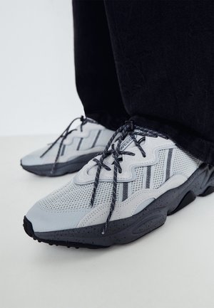 Baskets en mesh gris clair et noir avec des lacets à motifs, portées avec un pantalon noir sur un sol clair.