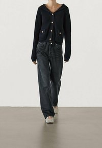 Granatowy kardigan z prążkowanej tkaniny, z kapturem i przednimi kieszeniami, nałożony na czarne jeansy o luźnym kroju, w zestawieniu z jasnoszarymi butami wsuwanymi.