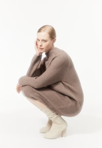Braunes gestricktes Kleid mit hohem Kragen und gerippter Textur, kombiniert mit cremfarbenen Stricksocken und strukturierten Absatzstiefeln. Model in kniender Pose.