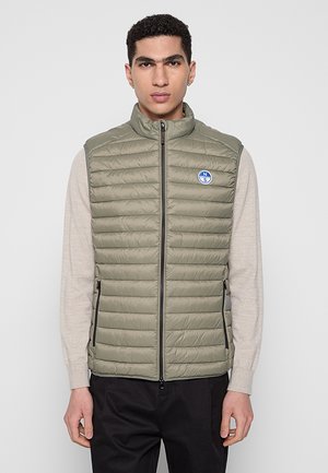 Uomo che indossa un gilet imbottito senza maniche color oliva chiaro con zip frontale e tasche laterali con zip sopra una camicia beige a maniche lunghe e pantaloni neri.