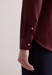 Bordeaux lange mouw shirt met een gladde textuur, voorzien van witte knopen en een geborduurd embleem op de manchet, schuin bij de pols.