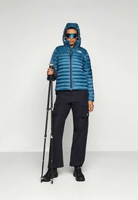 Puhasta tamnoplava izolirana jakna s kapuljačom, uparena s crnim cargo hlačama i crnim čizmama. Model drži štapove za planinarenje i nosi sunčane naočale.