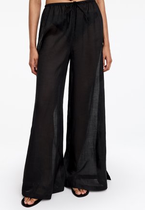 Pantalon noir transparent à jambes larges avec taille élastique et cordon, porté avec des chaussures noires ouvertes à l'avant.