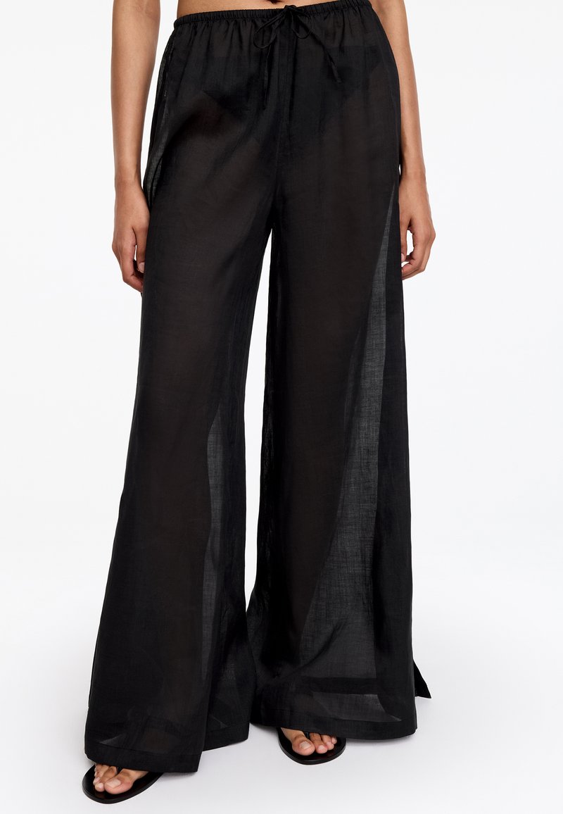 Pantalon noir transparent à jambes larges avec taille élastique et cordon, porté avec des chaussures noires ouvertes à l'avant.