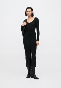 Μαύρο πλεκτό φόρεμα με ρίγες, που διαθέτει κουμπιά μπροστά, μακριά μανίκια, μήκος μέχρι τη μέση της γάμπας και σκίσιμο μπροστά. Φορεμένο με μαύρες μπότες μέχρι το γόνατο.