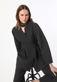 Blazer nero avvolgente con maniche lunghe, dotato di cintura in vita e pieghe. Indossato con una sciarpa a fantasia verde e rossa. Texture del tessuto liscia.