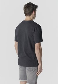 Man standing facing away wearing a black short-sleeve t-shirt and gray rolled-up denim shorts against a plain background.