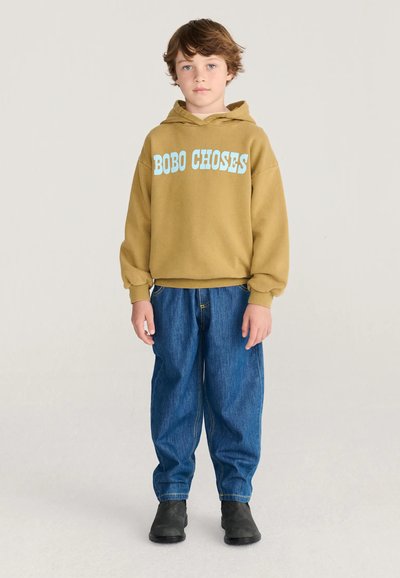 Enfant aux cheveux courts châtains portant un sweat à capuche moutarde avec le texte "BOBO CHOSES", un jean bleu ample et des chaussures noires, debout devant un fond uni.