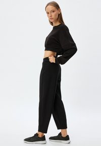 Sort cropped sweatshirt med lange ærmer, parret med højtaljede sorte brede bukser. Model iført sorte sneakers. Glat stofstruktur.