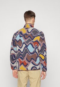 Fleece takki, jossa on värikäs aaltoileva kuvio ruskeassa, sinisessä, keltaisessa ja punaisessa. Hiukan kohotettu kaulus ja pitkät hihat.