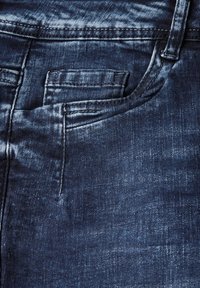Detailní pohled na tmavě modrou denimu, který ukazuje kapsu, švy a vybledlé oblasti, zvýrazňující texturu a barvu džínů.