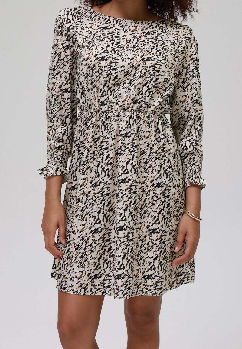 Femme portant une robe longue à manches longues jusqu'aux genoux avec des taches abstraites noires et marron sur fond blanc, et un bracelet en or à son poignet droit.