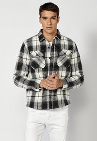 Zwart-witte geruite flanellen shirt met twee voorzakken, knoopsluiting en standaardkraag, gecombineerd met witte versleten jeans.