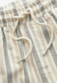 Gros plan d'un tissu rayé beige, gris et blanc avec une taille élastique et un cordon torsadé crème avec des embouts en métal.
