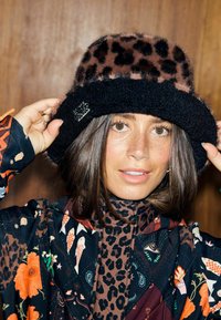 Pălărie de tip bucket cu imprimeu leopard maro și negru, cu textură pufoasă și margine din țesătură neagră. Are un accesoriu argintiu pe lateral.