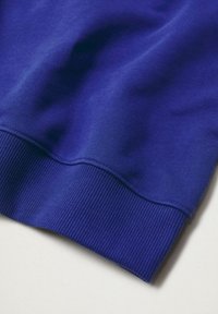 Gros plan sur le poignet d'une manche de sweat-shirt bleu roi avec une texture en maille côtelée visible sur fond blanc.