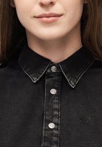 Κοντινό πλάνο στο κάτω μέρος του προσώπου και στον λαιμό ενός ατόμου που φοράει μαύρο τζιν πουκάμισο με ασημένια κουμπιά και έχει μακριά καστανά ίσια μαλλιά.