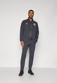 Sporting tracksuit in dark grey, features red accents and gold branding. Includes a zip-up jacket and tapered pants, made from lightweight fabric.
