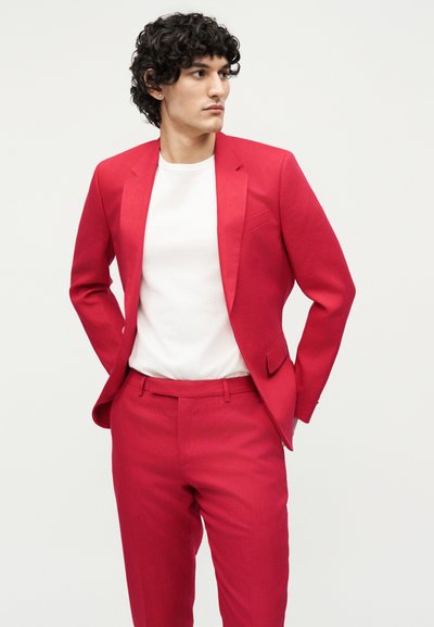 Άνδρας με κατσαρά μαλλιά που φοράει φωτεινό κόκκινο κοστούμι και λευκό Τ-shirt, με τα χέρια στις τσέπες, κοιτάζει προς τα πλάγια σε απλό φόντο.