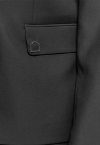 Tasca della giacca nera con una piccola spilla rettangolare d'argento a forma di arco attaccata alla patta.