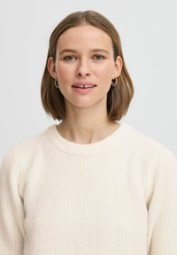 Pull en côtes crème avec un col rond, texture douce et manches longues. Un mannequin porte des boucles d'oreilles en or et un maquillage minimal.