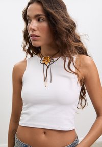 Bela brezrokavnik s kratkimi hlačami in gladko teksturo, kombinirana z rjavim vrvnim ogrlico z rumenim obeskom v obliki cvetlice in zlatimi kovinskimi poudarki.