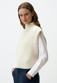 Pull en beige clair avec un col montant côtelé et des emmanchures larges, porté sur une chemise blanche à boutons, présentant un motif en maille texturée.