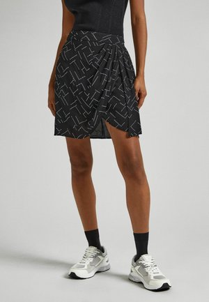Vrouw draagt een zwarte minirok met patroon, zwarte top, zwarte sokken en witte sportschoenen, staand op een effen lichte achtergrond.