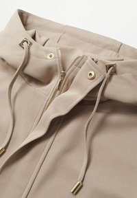 Chaqueta beige de cremallera con tejido texturizado, que cuenta con una capucha, detalles en dorado y cordones ajustables en el cuello.